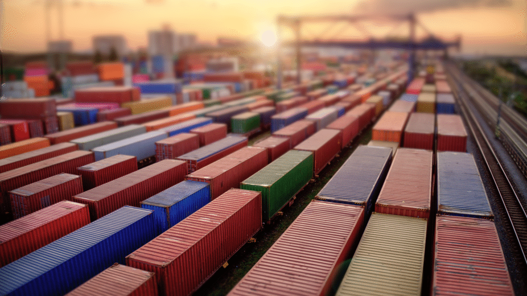 Luftaufnahme von Reihen bunter Schiffscontainer, die in einem großen Güterbahnhof bei Sonnenuntergang gestapelt sind, mit Kränen und Gleisen im Hintergrund.