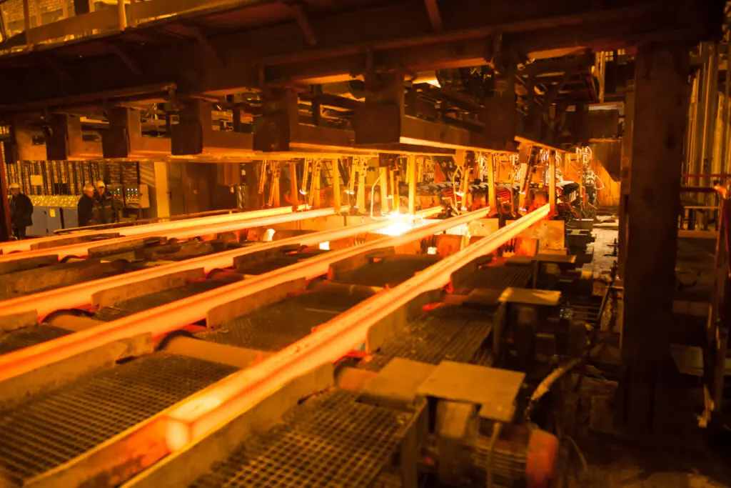 Reihen glühender Stahlträger bewegen sich auf Rollen in einem industriellen Stahlwerk, umgeben von schweren Maschinen und einem warmen orangefarbenen Licht, das den Innenraum der Fabrik erfüllt.