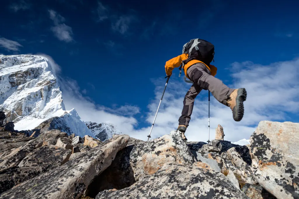 Ein Wanderer mit orangefarbener Jacke und Rucksack klettert mit Trekkingstöcken über felsiges Gelände, vor dem Hintergrund schneebedeckter Berge und eines strahlend blauen Himmels.