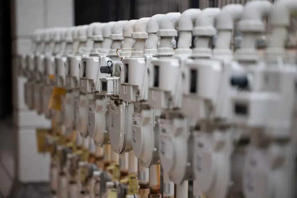 Eine Reihe von Industriegaszählern mit Rohren und Messgeräten, die nebeneinander an einer Wand aufgereiht sind und ein sich wiederholendes Muster bilden. Das Bild ist auf die Zähler im Vordergrund fokussiert, der Hintergrund ist unscharf.