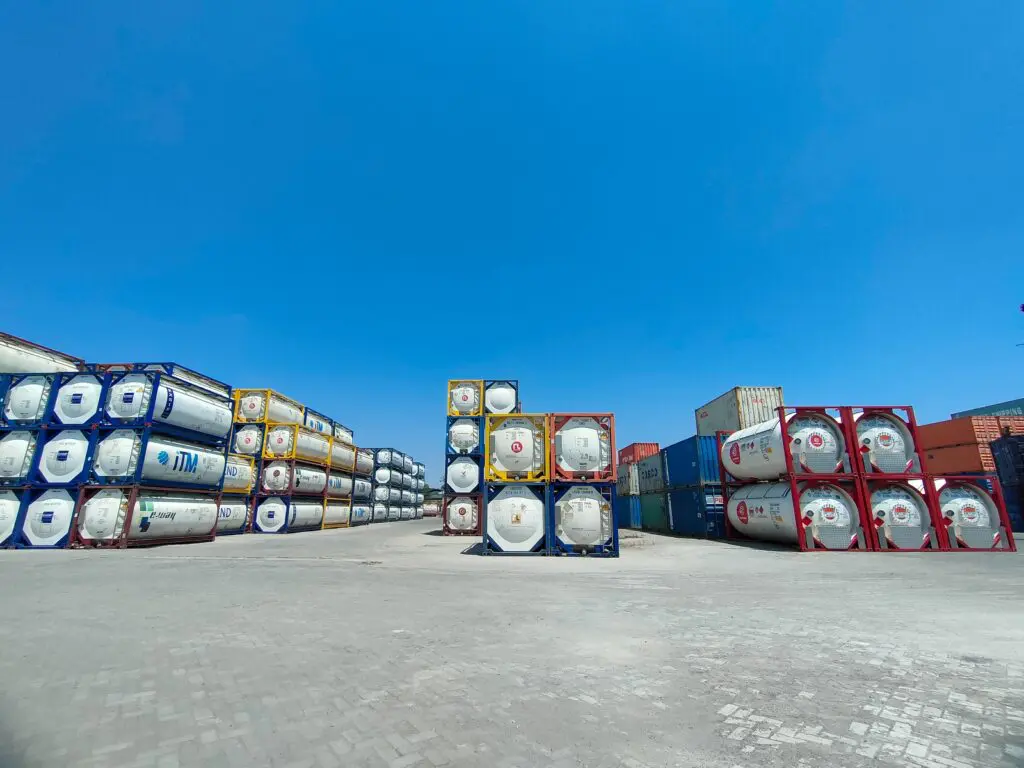 Auf einem gepflasterten Platz unter blauem Himmel sind zylindrische Tanks in Reihen angeordnet, im Hintergrund sind einige rechteckige Schiffscontainer zu sehen.