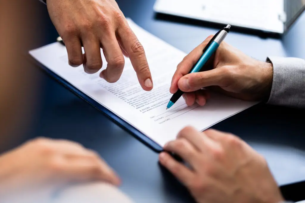 Nahaufnahme von zwei Personen, die ein Dokument prüfen; eine Person zeigt auf das Papier, während die andere einen Stift in der Hand hält und sich scheinbar auf die Unterzeichnung vorbereitet. Die Szene suggeriert eine Vereinbarung oder eine Vertragsunterzeichnung.