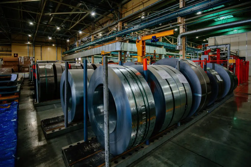 In einer geräumigen Industriehalle werden große Blechrollen aufrecht in Regalen gelagert, im Hintergrund sind Rohre und Geräte zu sehen.