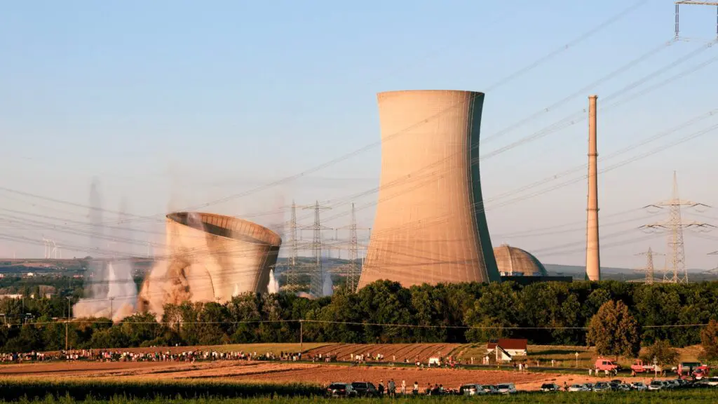 Ein Kühlturm eines Kraftwerks wird abgerissen und stürzt teilweise in den Staub ein, während andere Türme und Schornsteine in der Nähe stehen. Von einem Feld im Vordergrund aus schauen Menschen bei klarem Himmel zu.