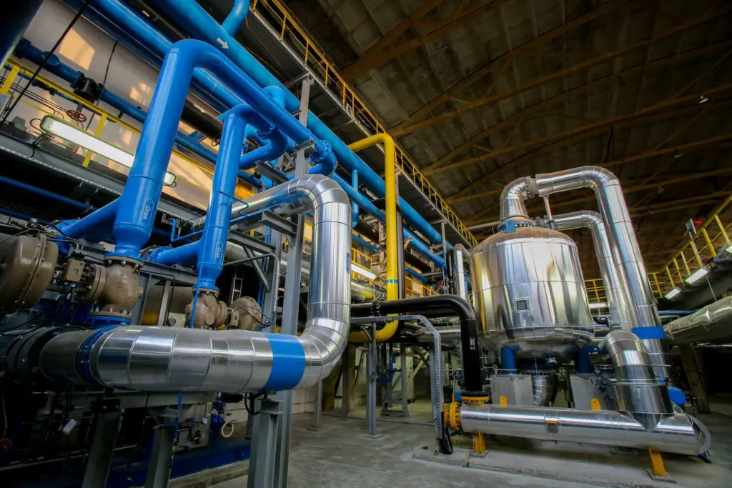 Das Innere einer Industrieanlage mit großen blauen, gelben und silbernen Rohren, die mit einem zylindrischen Metalltank verbunden sind, unter einer hohen Decke mit freiliegenden Balken. Der Bereich erscheint sauber und gut beleuchtet.