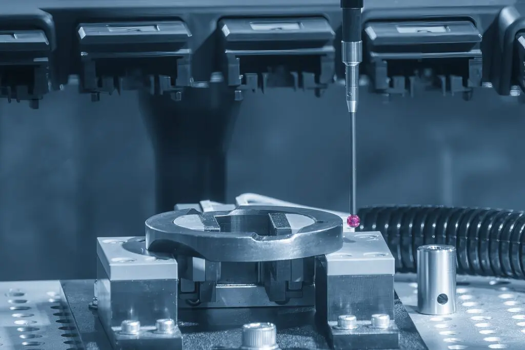 Nahaufnahme eines Koordinatenmessgeräts (KMG), das ein Metallteil auf einem industriellen Prüftisch prüft, wobei im Hintergrund Präzisionsgeräte und Maschinenteile zu sehen sind.