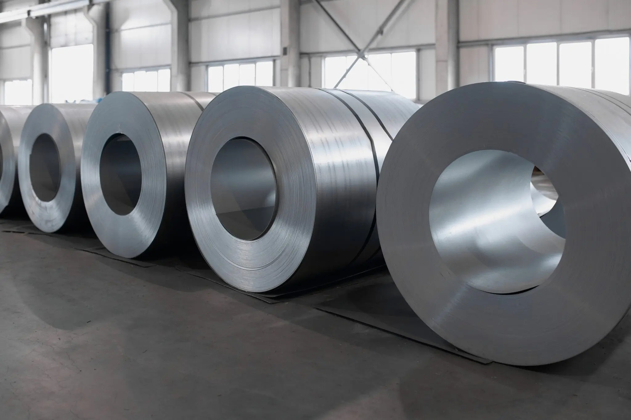 Große Rollen glänzenden Stahlblechs liegen auf dem Boden eines hellen Industrielagers aufgereiht, bereit zur Verarbeitung oder zum Transport.