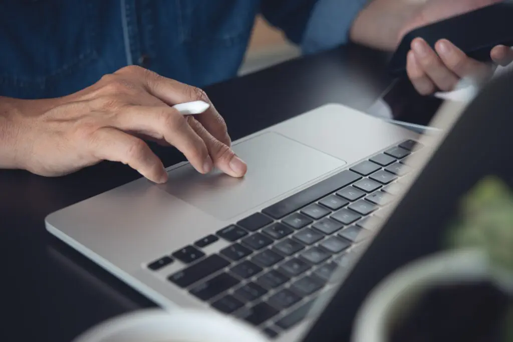 Eine Person, die einen Laptop benutzt, eine Hand auf dem Trackpad und die andere hält ein Smartphone, arbeitet an einem Schreibtisch mit einer verschwommenen Kaffeetasse und einer Pflanze im Vordergrund.