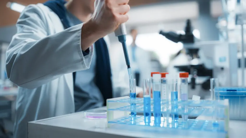 Ein Wissenschaftler in einem Laborkittel benutzt eine Pipette, um Flüssigkeit in Reagenzgläser auf einem Labortisch zu füllen. Im Hintergrund sind verschiedene Laborgeräte und ein Mikroskop zu sehen.