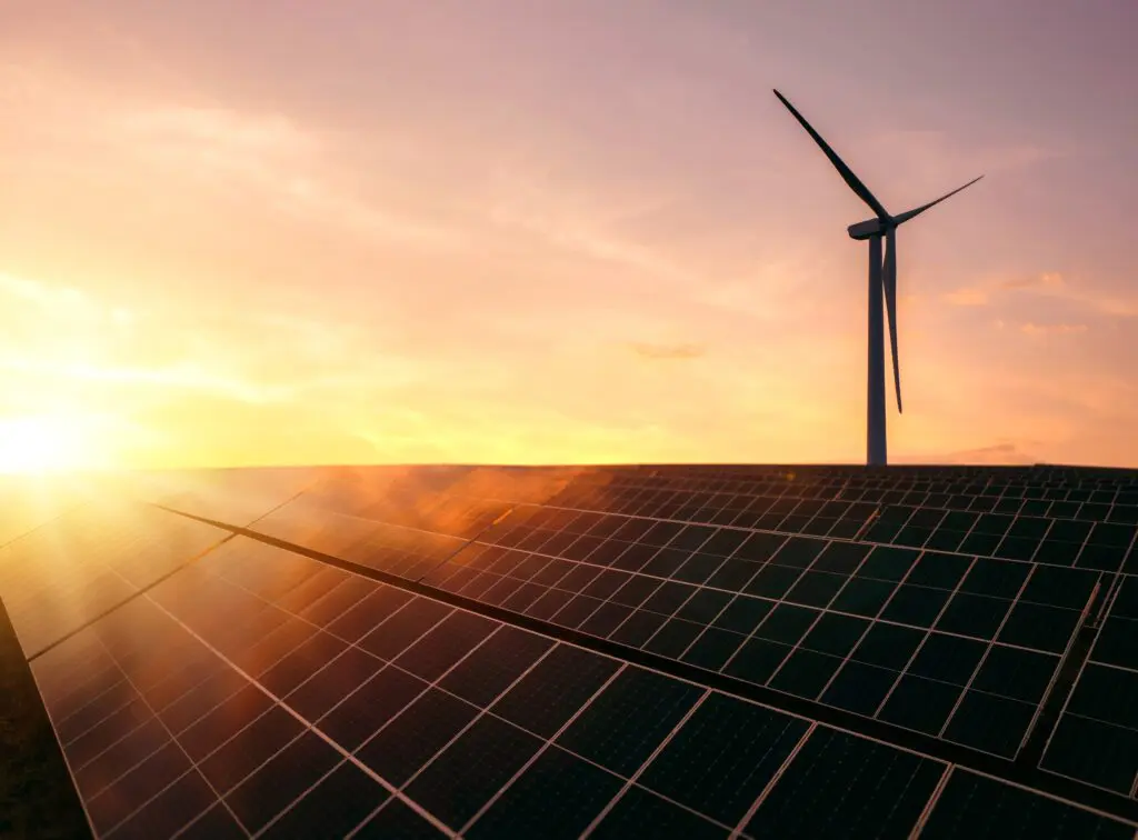 Sonnenkollektoren und eine Windturbine zeichnen sich vor einem leuchtend orange-gelben Sonnenuntergang ab und symbolisieren erneuerbare Energiequellen.