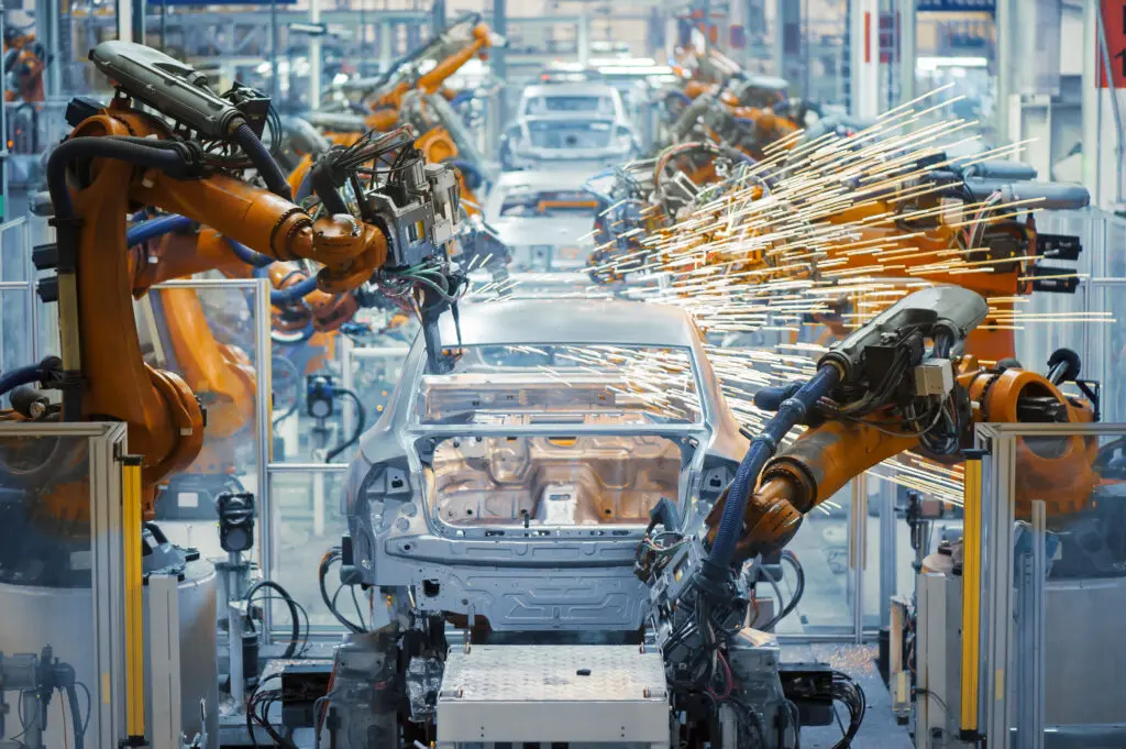 Roboterarme arbeiten an der Montage eines Fahrzeugrahmens und erzeugen dabei Funken in einer hell erleuchteten, modernen Automobilfabrik mit mehreren Fahrzeugen in verschiedenen Montagestufen.