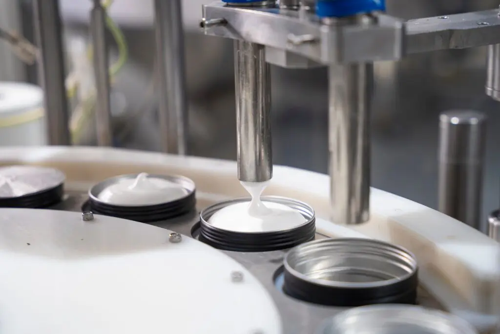 Nahaufnahme einer Maschine, die weiße Sahne in offene Gläser mit schwarzem Deckel auf einer Fertigungsstraße in einer Fabrikumgebung ausgibt.