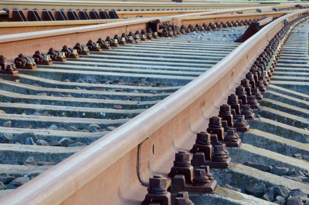 Nahaufnahme von rostigen Eisenbahnschienen und Metallbefestigungen mit mehreren parallel verlaufenden und leicht divergierenden Schienensträngen, die auf Schotter und Betonschwellen ruhen.
