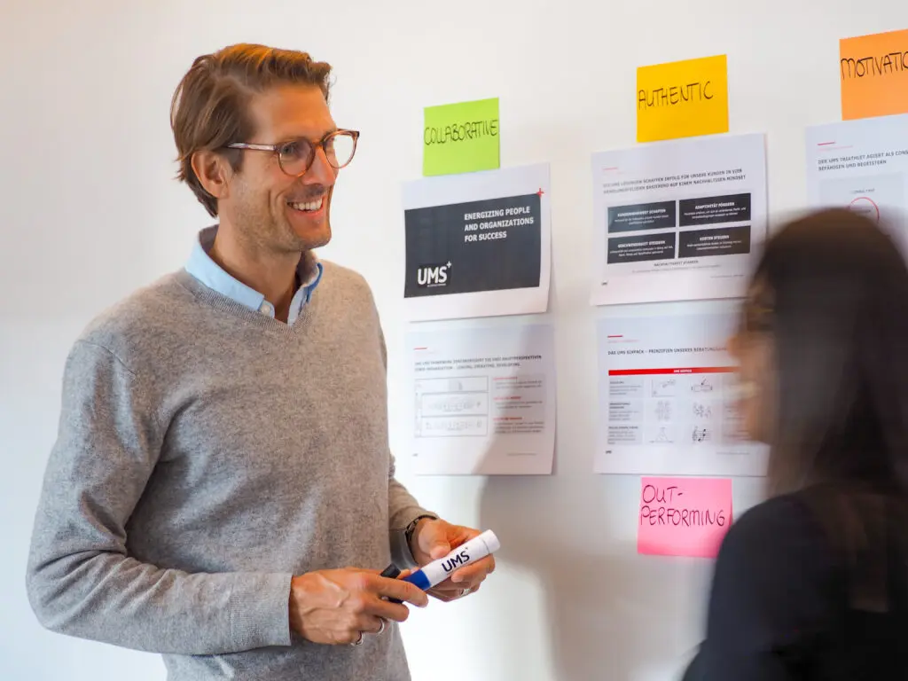 Ein Mann mit Brille und grauem Pullover lächelt, während er einen Marker in der Hand hält. Er steht an einer Wand mit Geschäftspapieren und bunten Etiketten wie Collaborative und Authentic. Eine Frau steht in der Nähe, leicht unscharf.