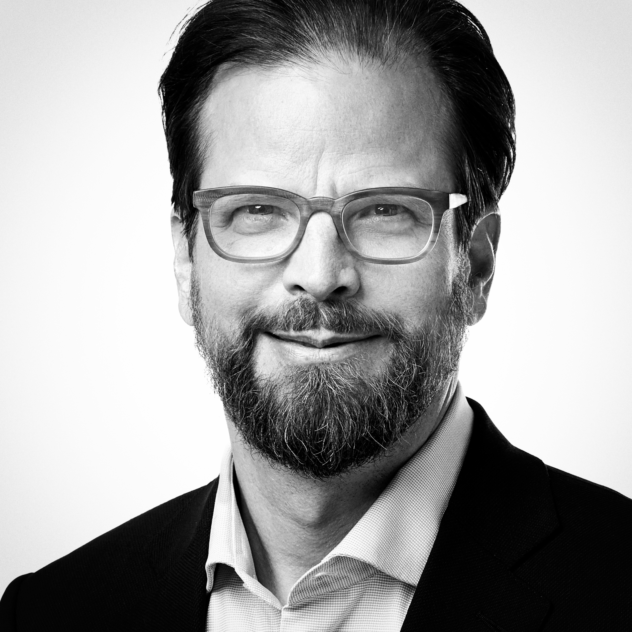 Schwarz-weißes Porträt eines Mannes mit Brille, Bart und Schnurrbart, der ein Hemd mit Kragen und einen Blazer trägt und leicht lächelt, vor einem schlichten hellen Hintergrund.