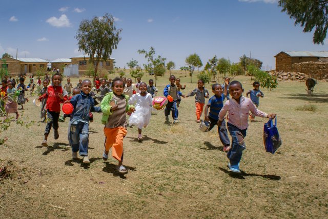 Eine große Gruppe lächelnder Kinder rennt mit Taschen und Behältern in der Hand energisch über eine trockene Wiese auf die Kamera zu, während im Hintergrund Gebäude und Bäume unter blauem Himmel zu sehen sind.
