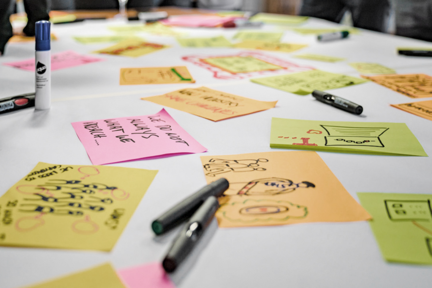 Auf einem weißen Tisch liegen bunte Haftnotizen mit Zeichnungen und handschriftlichen Ideen verstreut, umgeben von Markern ohne Kappe – ein Hinweis auf ein gemeinsames Brainstorming oder eine kreative Planungssitzung.